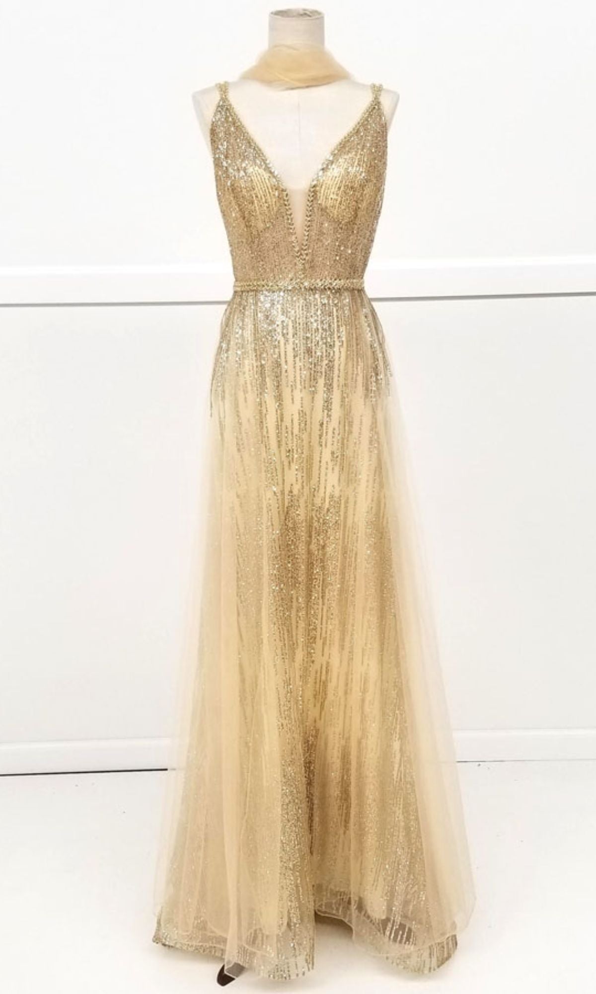 The Sheer-Waist Long Sequin Prom Dress: Chicas YG5021 is a gold, floor-length gown with a deep V-neckline, sequin detailing, and a sheer overlay, shown on a mannequin.